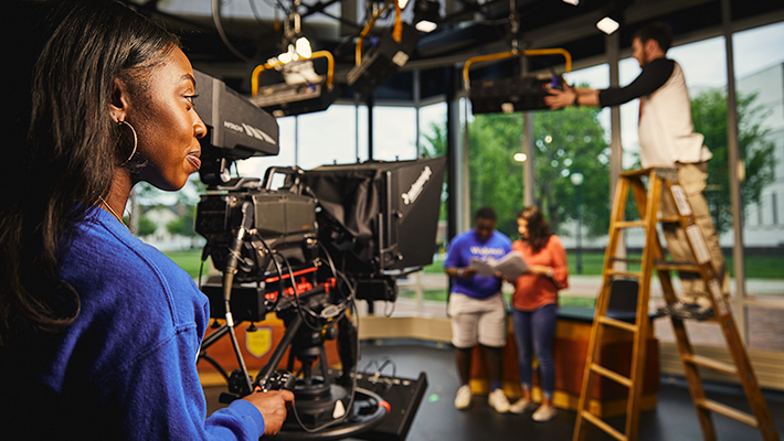 Broadcast student in studio 710x400 Broadcast commiunication student preparing to film in studio while classmates set up equipment