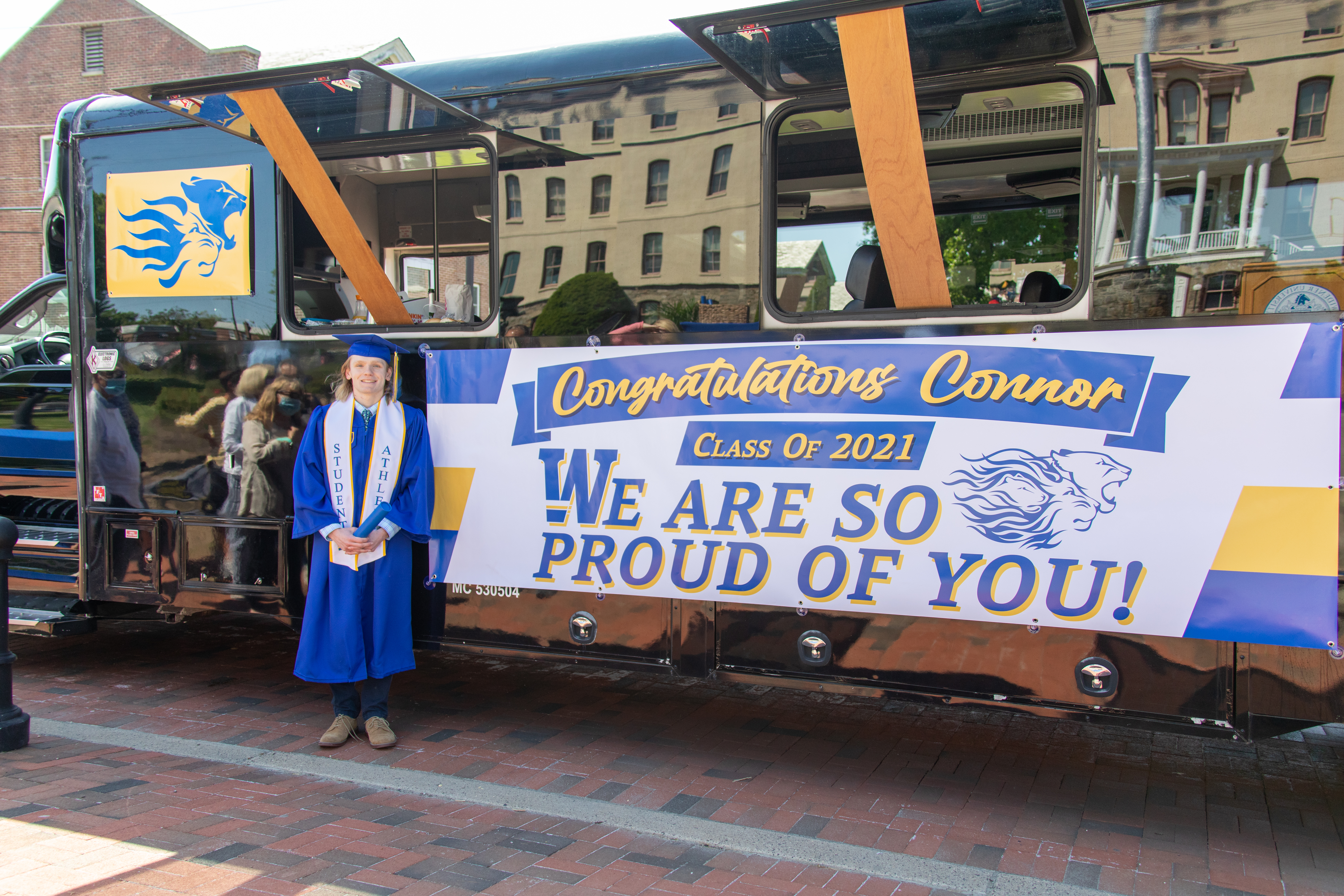 Graduate stands in front of black bus and sign that says "Congratulations Connor, Class of 2021, We are so proud of you!"