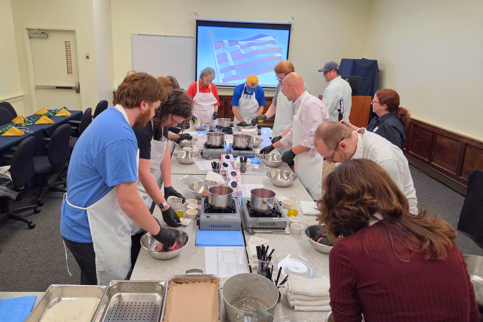 Students and staff stand around a long table with bowls and burners cooking a dish