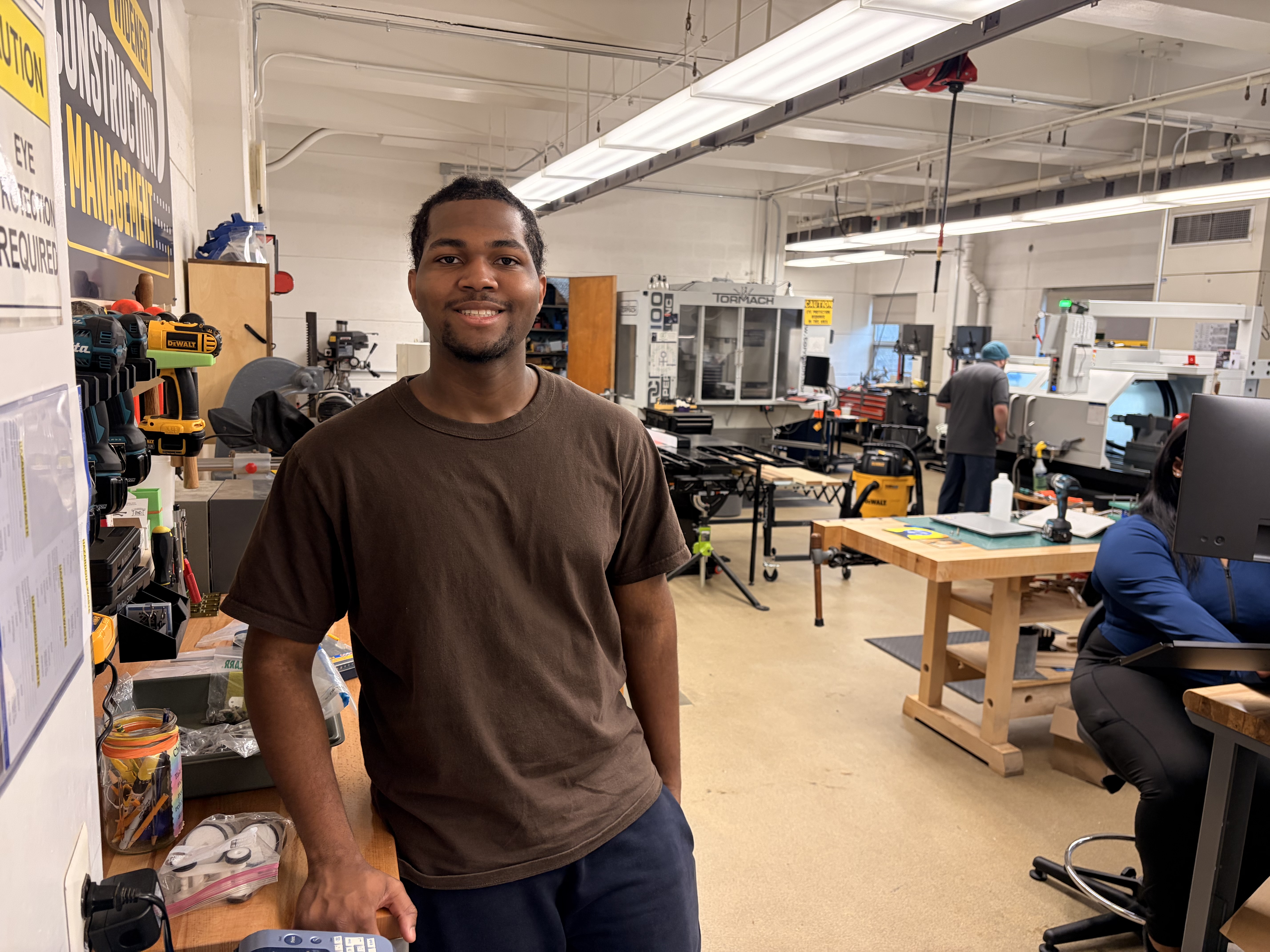 Clauddavid smiles with the various makerspace and machine shop equipment behind him.