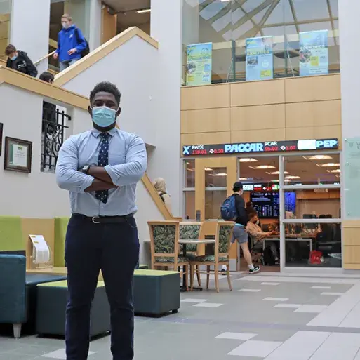 Brandon Matambo stands in the lobby of Quick Center; behind him is a stock ticker and students walking down steps