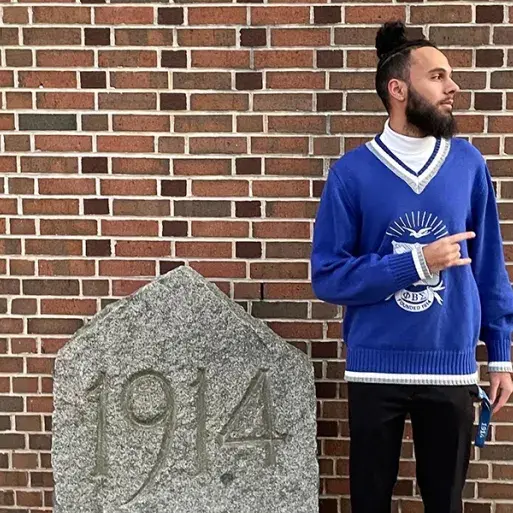 Adonijah Allsup, wearing a blue fraternity sweater, poses next to a stone marked 1914