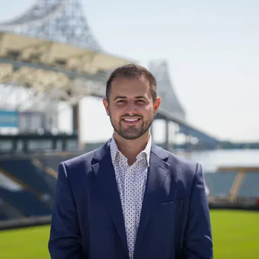 Niko Dinoulis wears a blue suit and poses in front of the union stadium overlooking a bridge and river