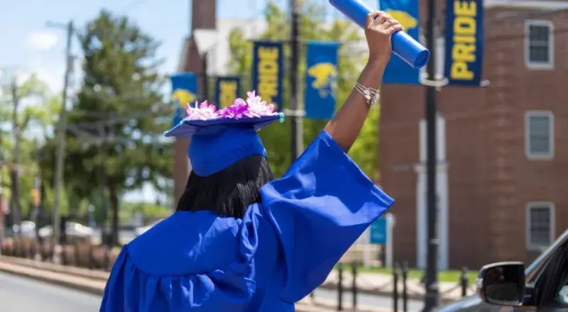 A female graduate in a cap and gown raises her diploma in the air.