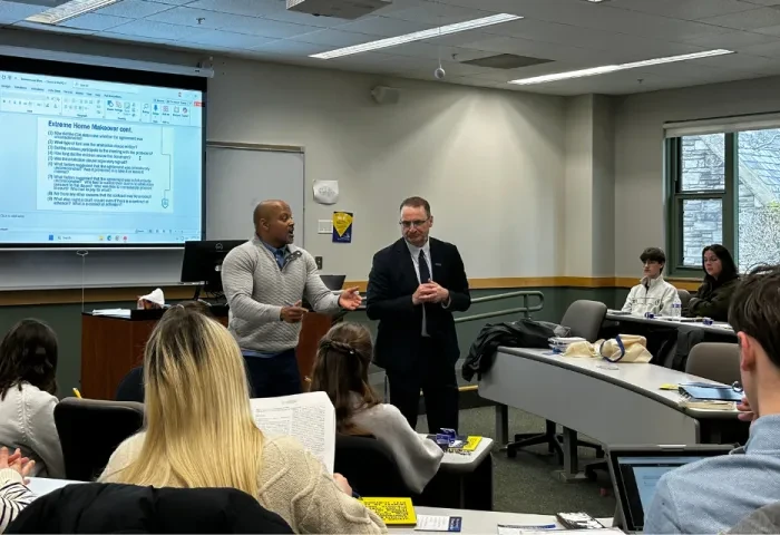 Deans Todd Clark and andré douglass pond cummings stand at the front of the room speaking to students. They have a presentation displayed on the screen behind them.