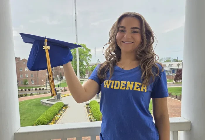 Amanda Rappa, wearing a blue Widener shirt and holding a blue grad cap, stands on the balcony of Old Main with campus behind her