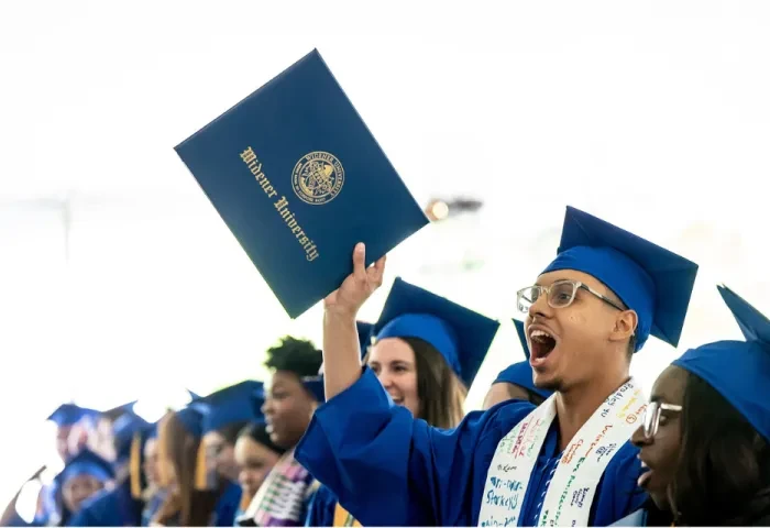 A student wearing a blue graduation cap and gown holds up his diploma case and cheers