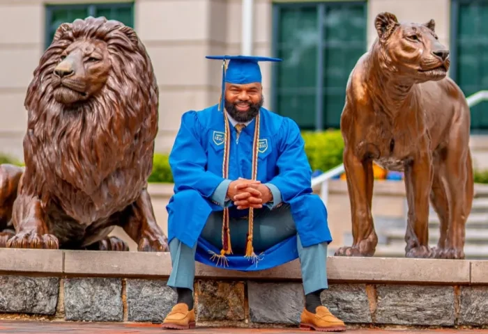 Delorean Andrews poses in his graduation cap and gown with the Pride of lions statue on campus.