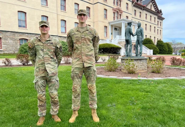 R-O-T-C cadets Liam Reilly and Braydn Erb, wearing Army fatigues, stand on the grass in front of Old Main with a statue in the background