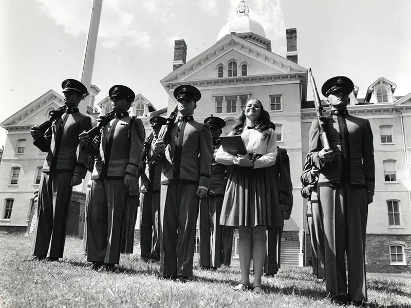 Black and white photo of a female student standing next to a group of cadets in front of Old Main