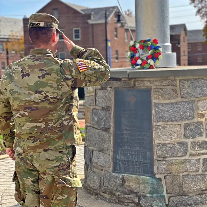 Back of an ROTC cadet in fatigues as they salute in front of a flag pole where a wreath rests