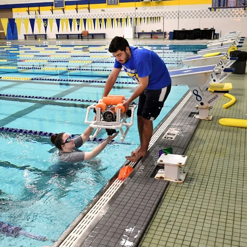 A male student helps lower an autonomous submarine into a pool to a female classmate to test the machine.