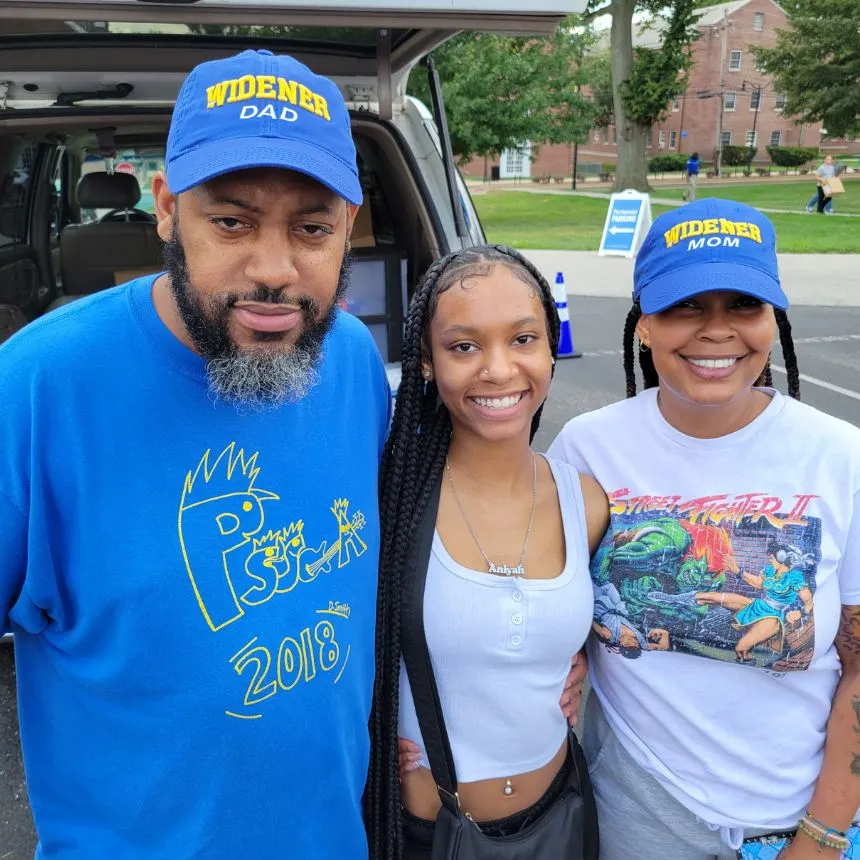 A female incoming student poses with her parents who are both wearing "Widener Dad" and "Widener Mom" baseball caps.