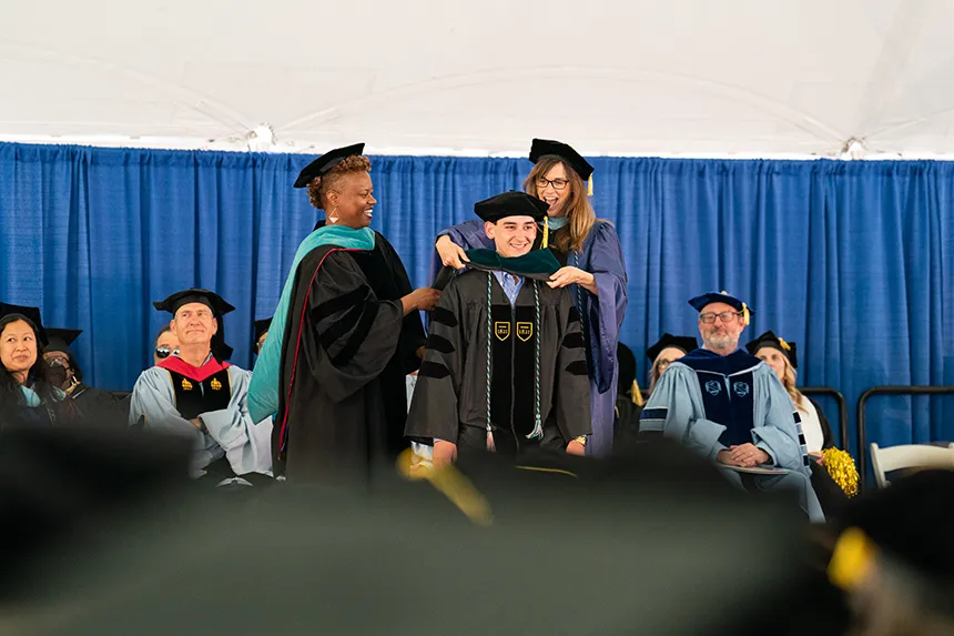 A physical therapy doctoral student has his hood placed over his head on stage