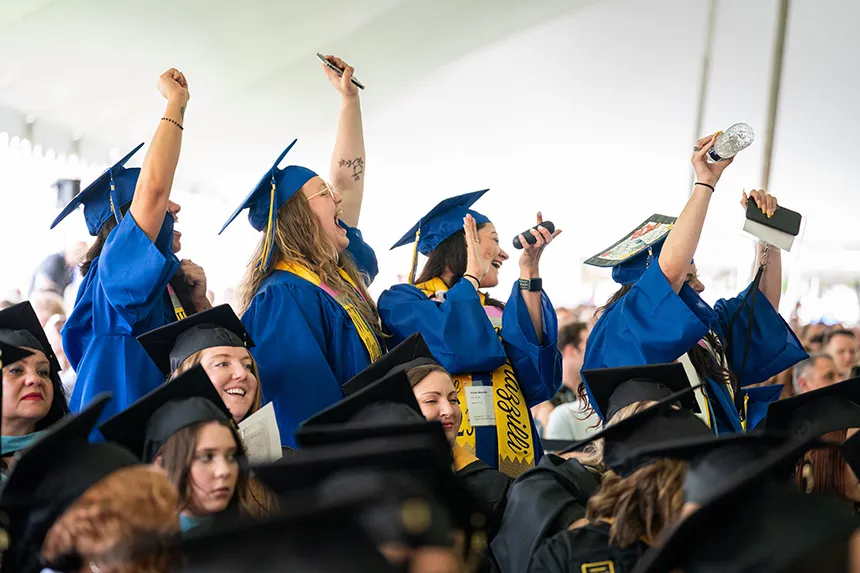 A group of grads in blue caps and gowns stand in their seats to cheer