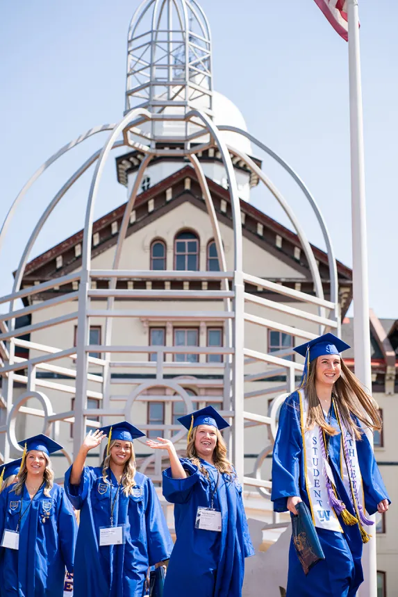 A group of grads in blue caps and gowns walk through the Bown Garden