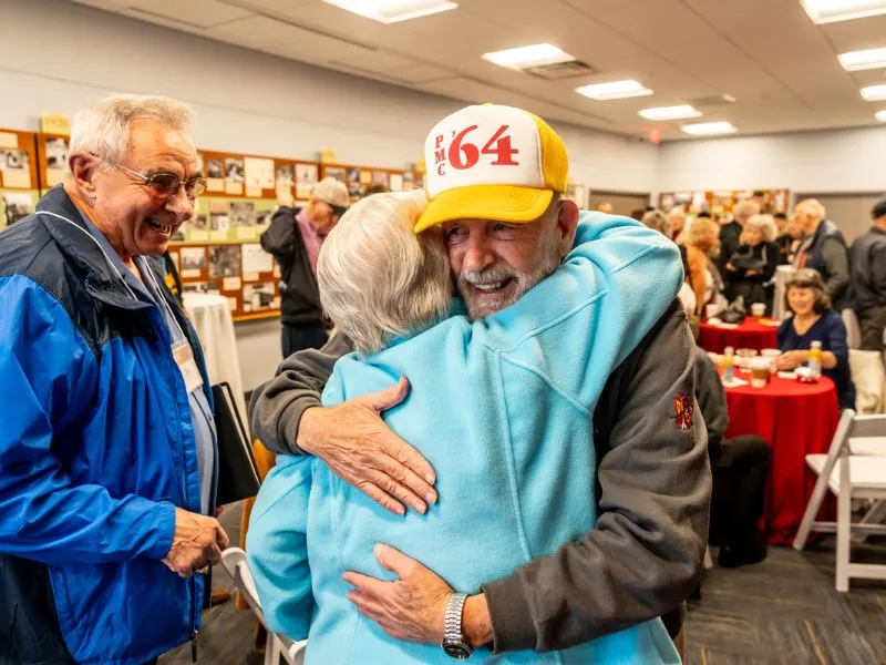 PMC alumni hug. One alumnus wears a '64 alumni hat.