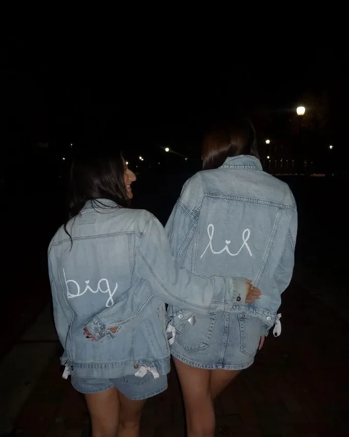 Two sorority members walking together at night wearing matching denim jackets labeled “big” and “lil.”