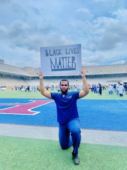 Alum Jeremiah McFarland, wearing nursing scrubs and kneeling while holding a Black Lives Matter sign