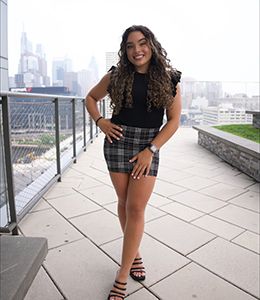 Student Trustee Maritza Garcia poses for a photo outdoors, with a city skyline behind her. city 