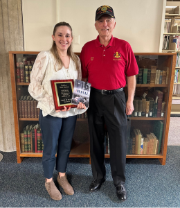 Kayla Van Osten and Ken Byerly post for a photo in front of a bookcase. Kayla holds Ken's book and a plaque.