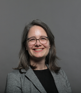 Lisa Grimm smiles at the camera in front of a gray background wearing a black top and gray jacket