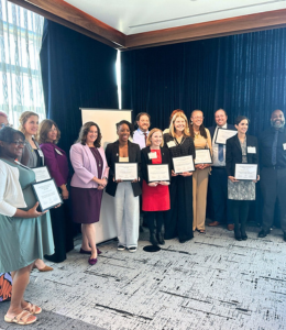 A group of people stand posing with their award papers, smiling.
