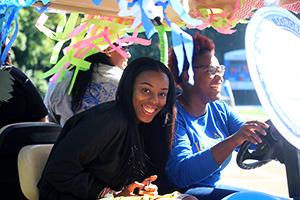 Student organizations golf cart parade 300x200 Two Widener students in a colorfully decorated golf cart driving on the track as part of the student organizations golf cart parade.