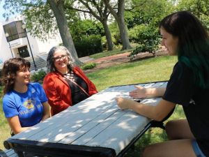 Student leaders, faculty member of Q&A Queer Zine sit at table outside Library 500x375 Two students and a faculty member sit at a picnic table with library in background