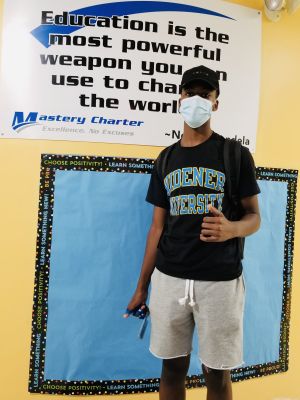 Peyton Brame, wearing a Widener T-shirt, stands in front of a sign at his high school, Mastery Charter