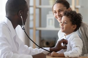 A male doctor uses a stethoscope on a young patient, who is held by her mother.