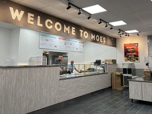 Interior of the Moe's location on campus with a sign that says &quot;Welcome to Moe's&quot; and two people working behind the counter