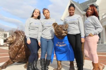 Four students in "We're All Widener" shirts stand with the Pride Lion statue