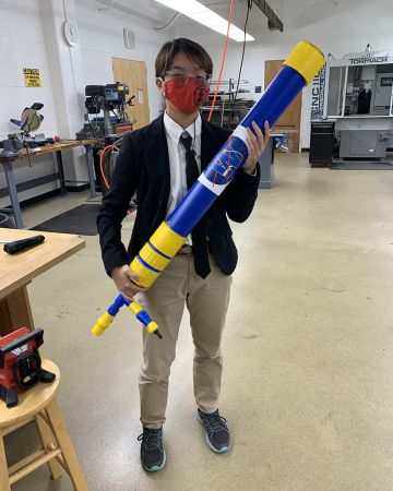 Engineering student stands in the machine shop holding a blue and yellow T-shirt cannon
