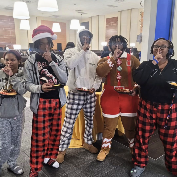 Five students in Christmas-themed pajamas hold plates of breakfast foods in the dining hall and hold a finger to their mouths in a silence gesture