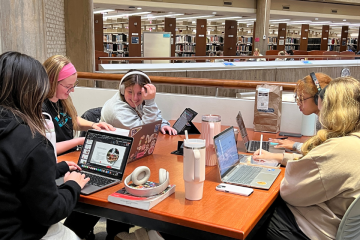 Five students sitting at a table in the library with open laptops and books