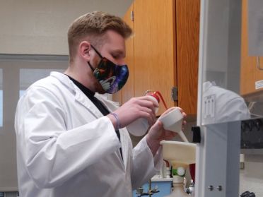 Aidan Looby, wearing a white lab coat and mask, works with equipment in a science lab