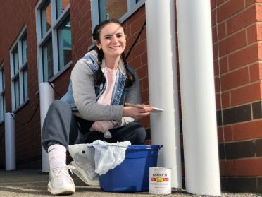 Christina Burke smiles and knees in front of a building while painting the exterior.