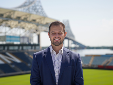 Niko Dinoulis wears a blue suit and poses in front of the union stadium overlooking a bridge and river