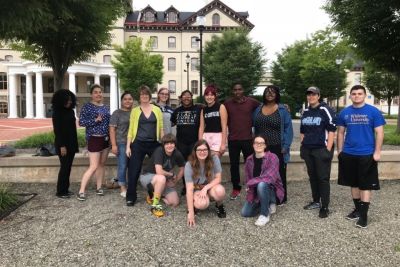 A group of 13 students and 1 professor pose in front of the Old Main administrative building