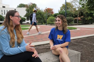 Two female students sit outside talking while other students walk past.