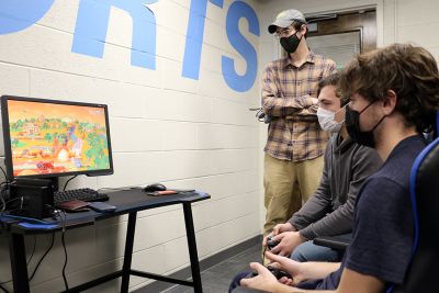 Two students play esports while Devin Hartnett stands next to them watching