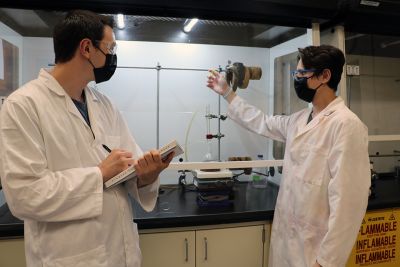 Two students in lab coats, masks and goggles work in a lab with equipment and a notebook