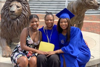 Three members of the McRae family sitting in front of the Pride of Lions statue, with one daughter in commencement regalia.