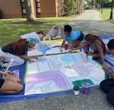 Group of students paint a piece of the mural on the ground on campus