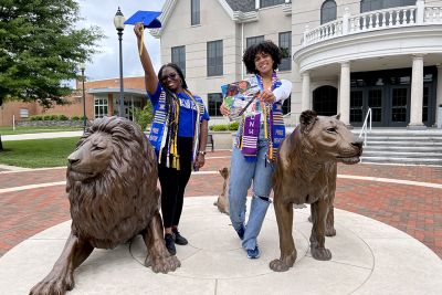 Two students in grad cords and stoles pose next to the Pride lion statues while holding their grad caps