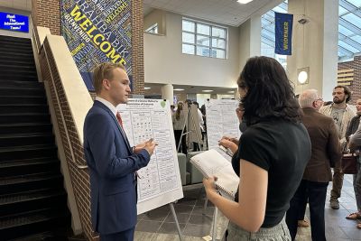 A student stands in front of a poster presentation speaking to another student who is taking notes.