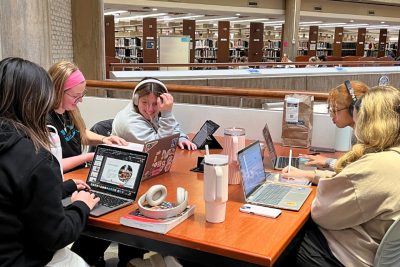 Five students sitting at a table in the library with open laptops and books
