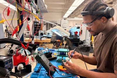 A young engineering student sits at work station in an engineering lab surrounded by equipment and machinery.