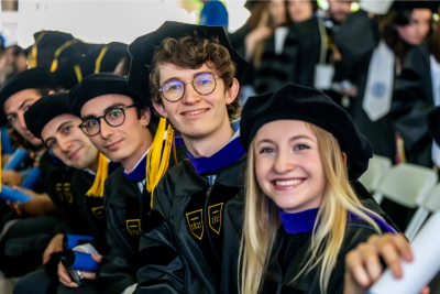 Graduating law students in their caps and gowns smile at the camera from their seats.